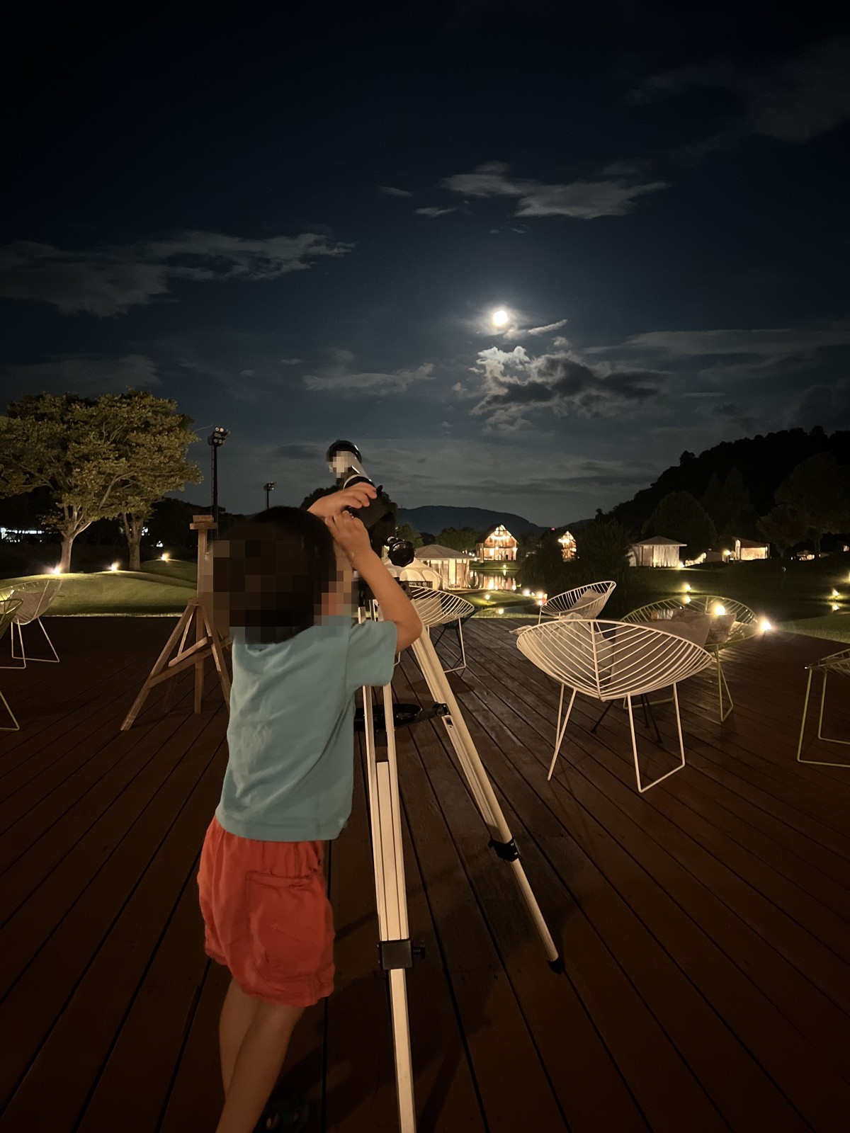 夜のデッキで望遠鏡をのぞく子ども。空には月