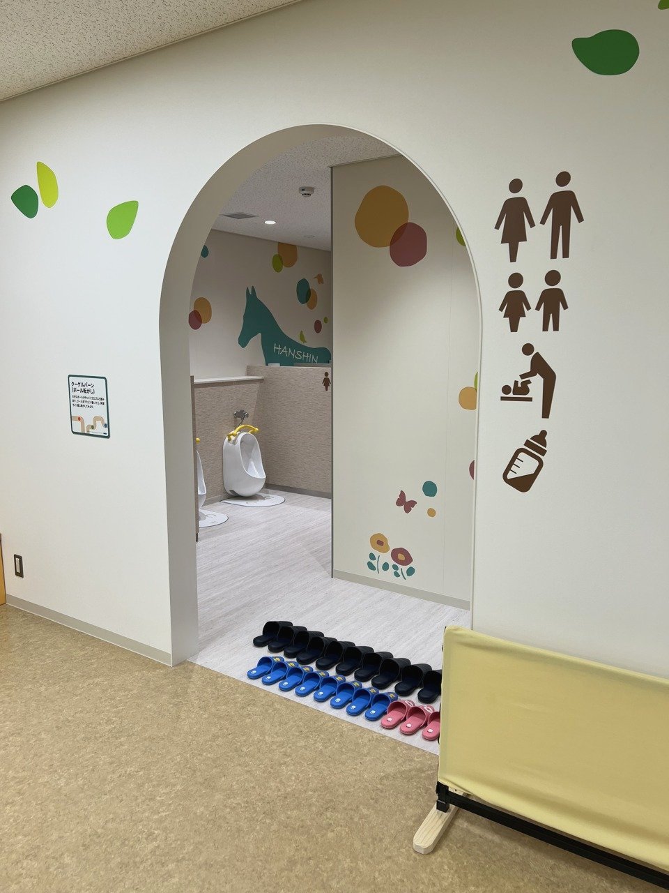 アーチ形の入口の奥に子ども用の小さい便器が並ぶトイレ。入口の横に大人・子ども・おむつ替え・ミルクのマーク