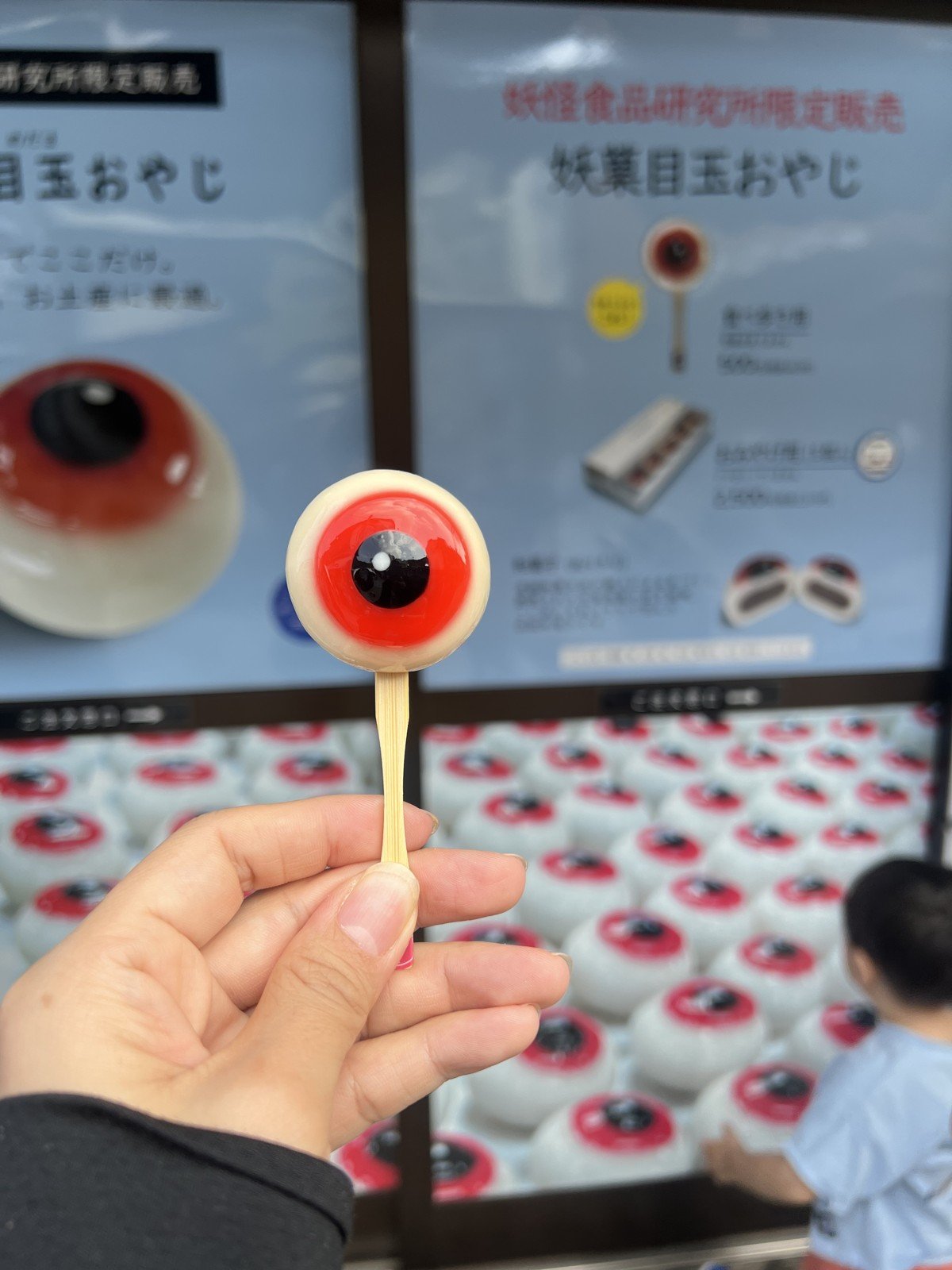 「妖菓目玉おやじ」を手に持つ。うしろに目玉のお菓子がずらり