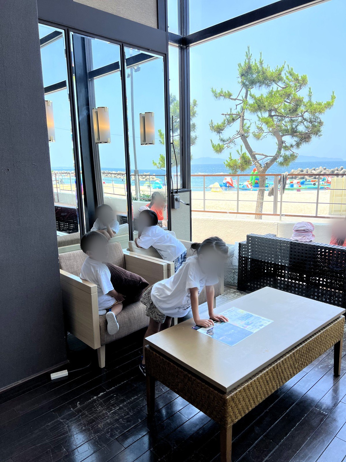 ロビーの窓際のソファでくつろぐ子どもたち。窓の外に砂浜と海上アスレチック