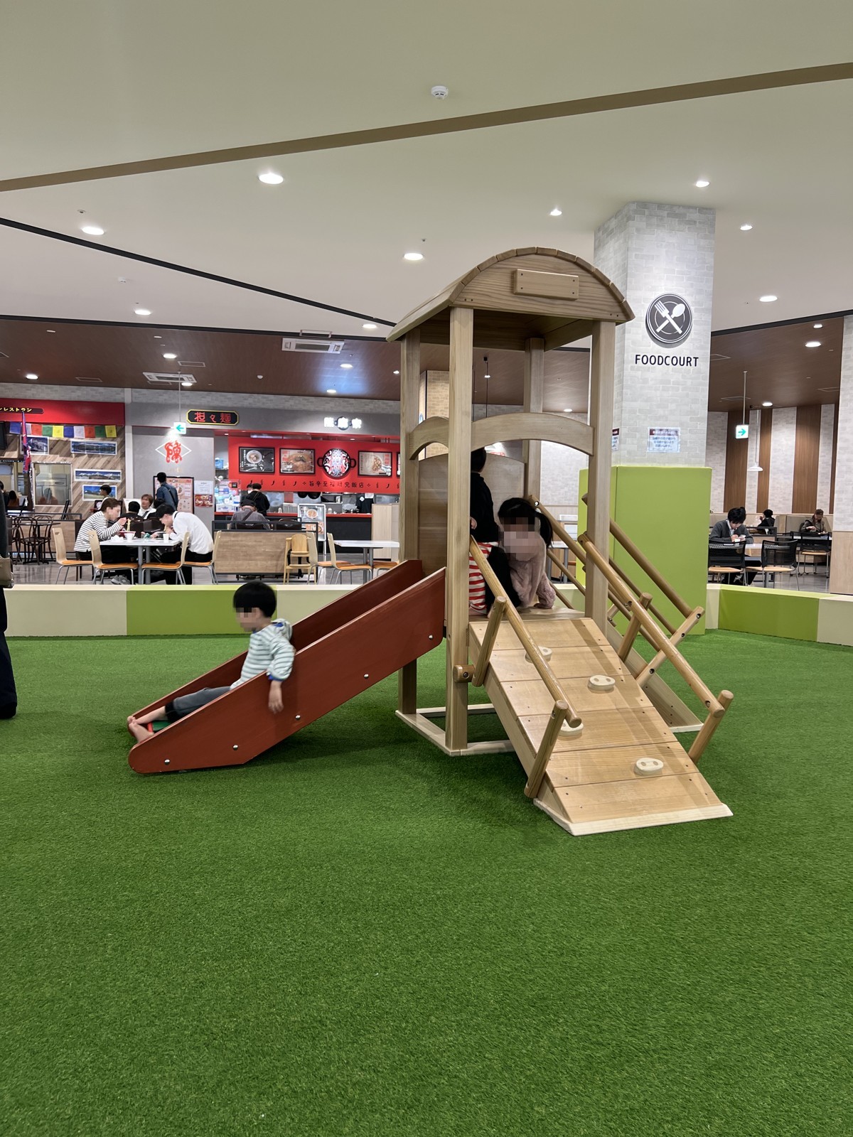 人工芝の上に置かれた木のすべり台つき遊具。奥はフードコート