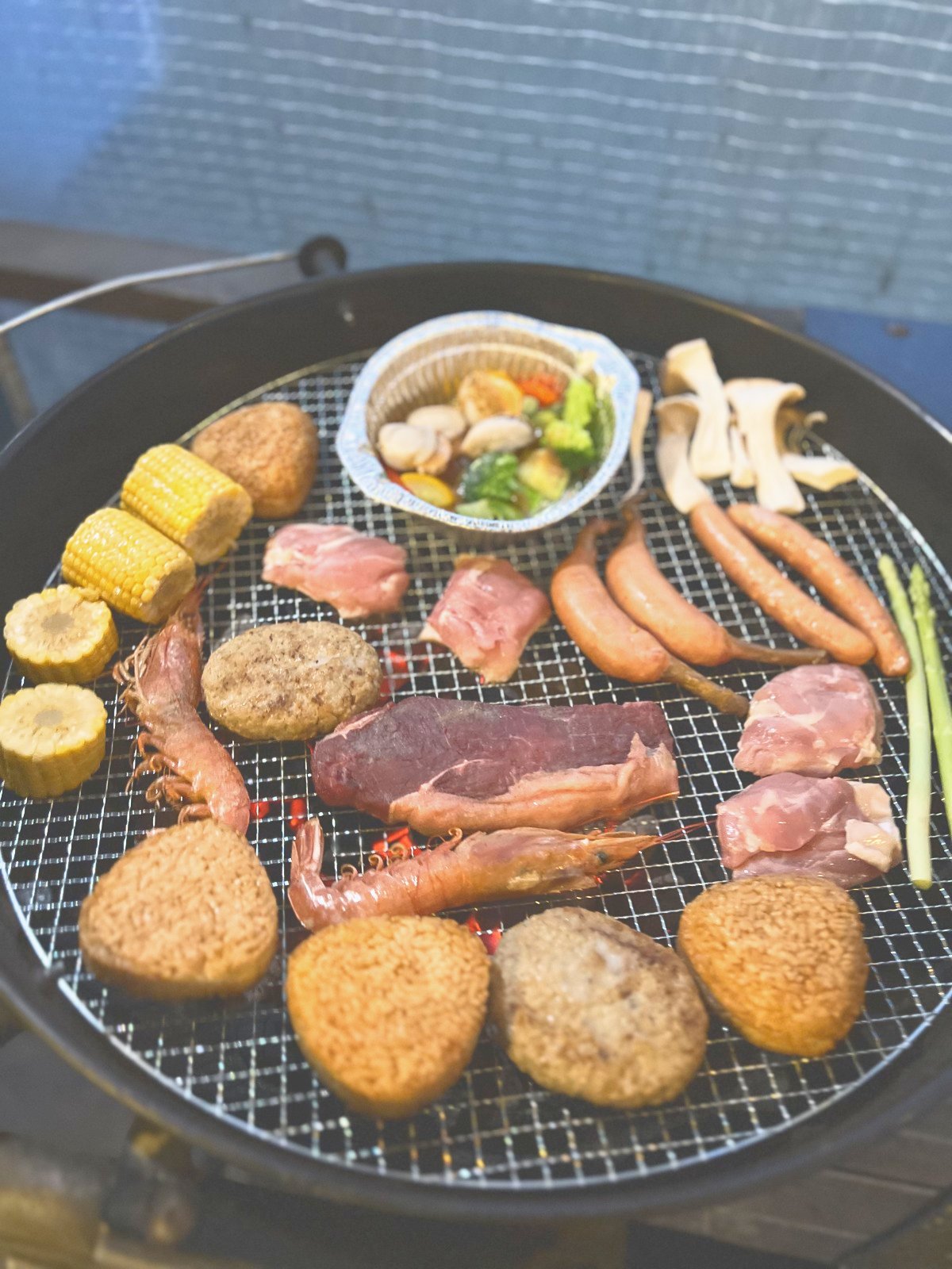 丸い炭火グリルに並ぶ肉・ソーセージ・とうもろこし・ハンバーグ