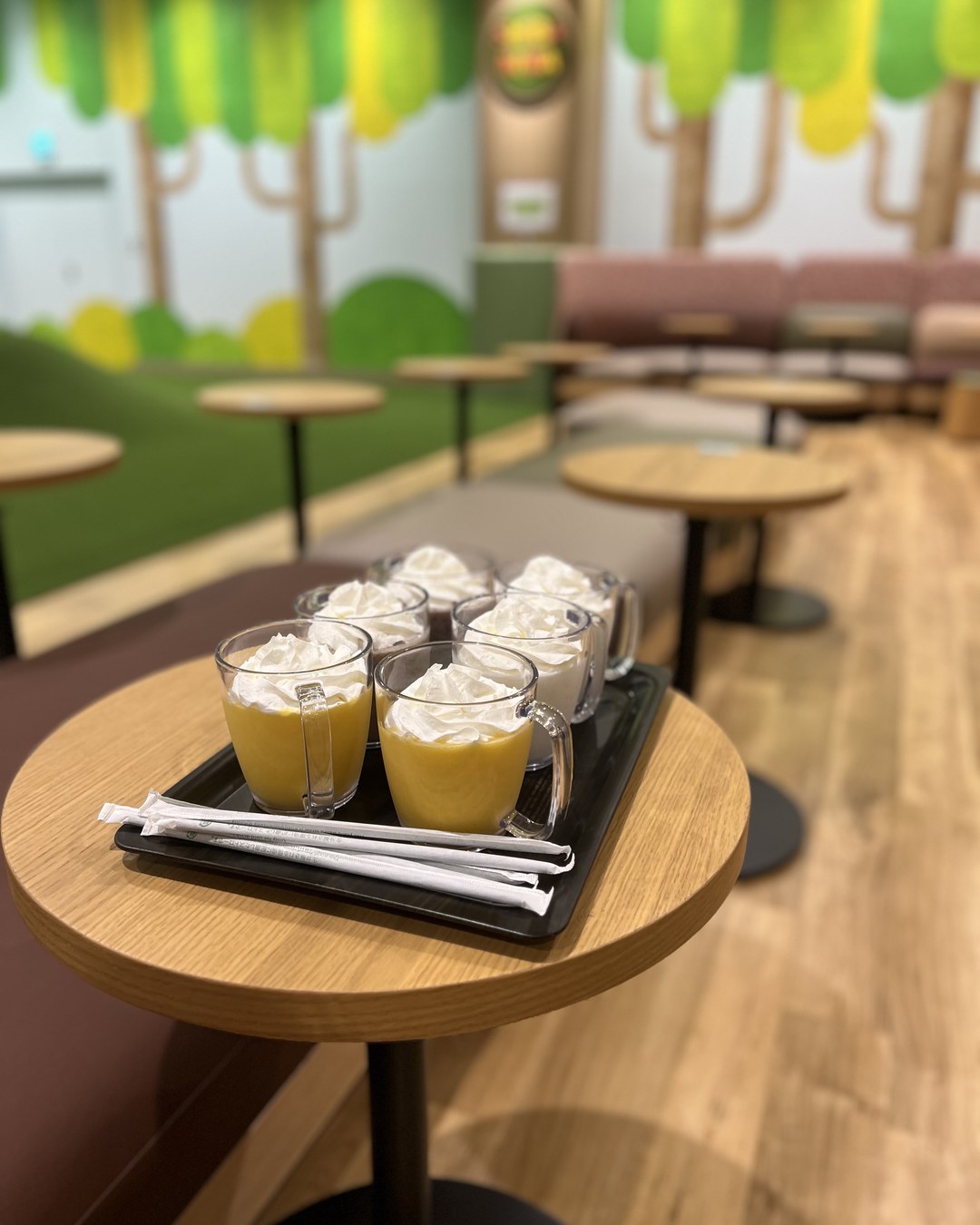 トレイにのった取っ手付きカップのドリンク4杯と、木の壁画のある店内